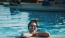 woman with sunglasses, in a pool, rests her arms on the ledge and smiles