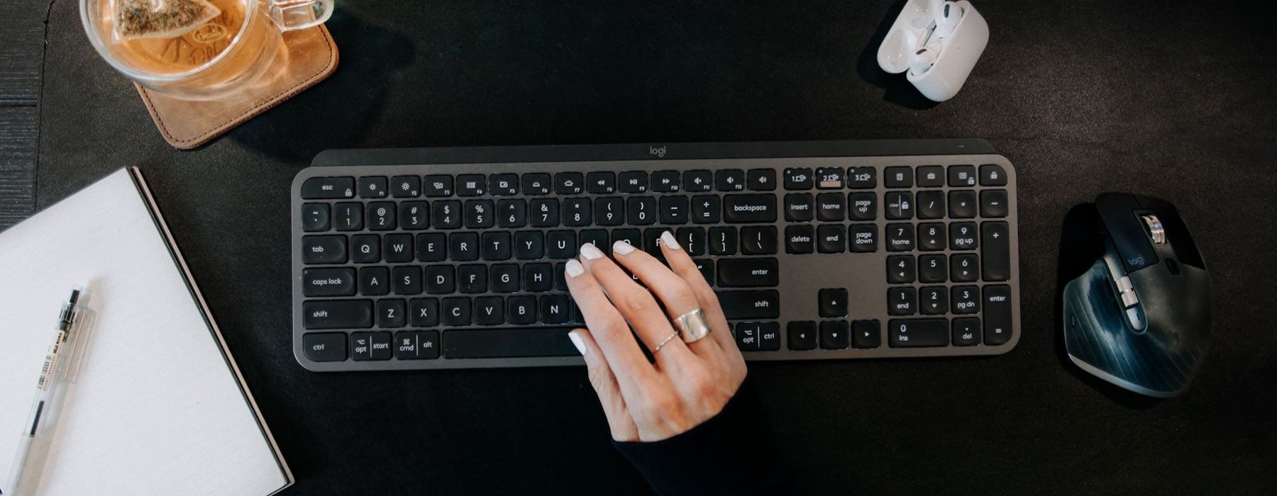 woman's hand on a keyboard surrounded by office items and a cup of tea on a coaster